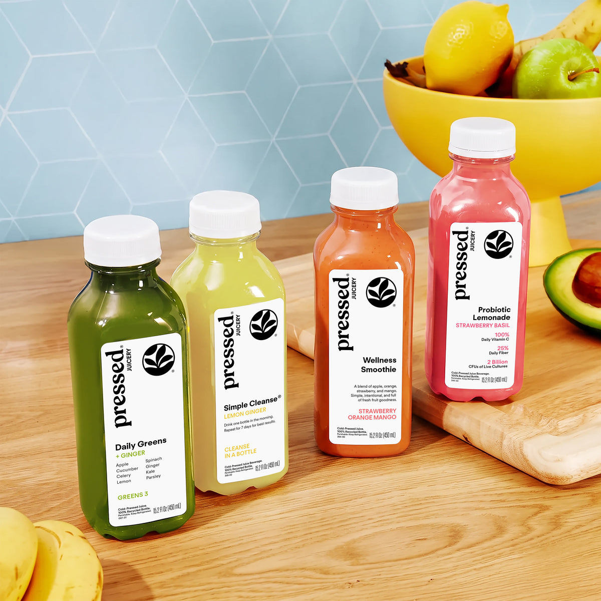 Four bottled juices from pressed brand arranged on a wooden countertop in a kitchen setting, with a bowl of fruit and a blue hexagonal backsplash in the background. The bottles are green, yellow, orange, and pink, labeled Daily Greens, Simple Cleaner, Wellness Smoothie, and Probiotic Lemonade.