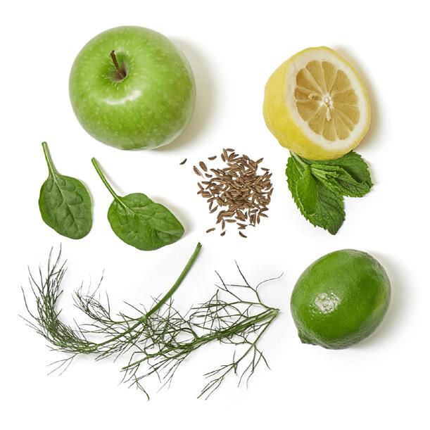 Collection of green fruits and vegetables including an apple, lemon, lime, spinach, dill, and cumin on a white background.